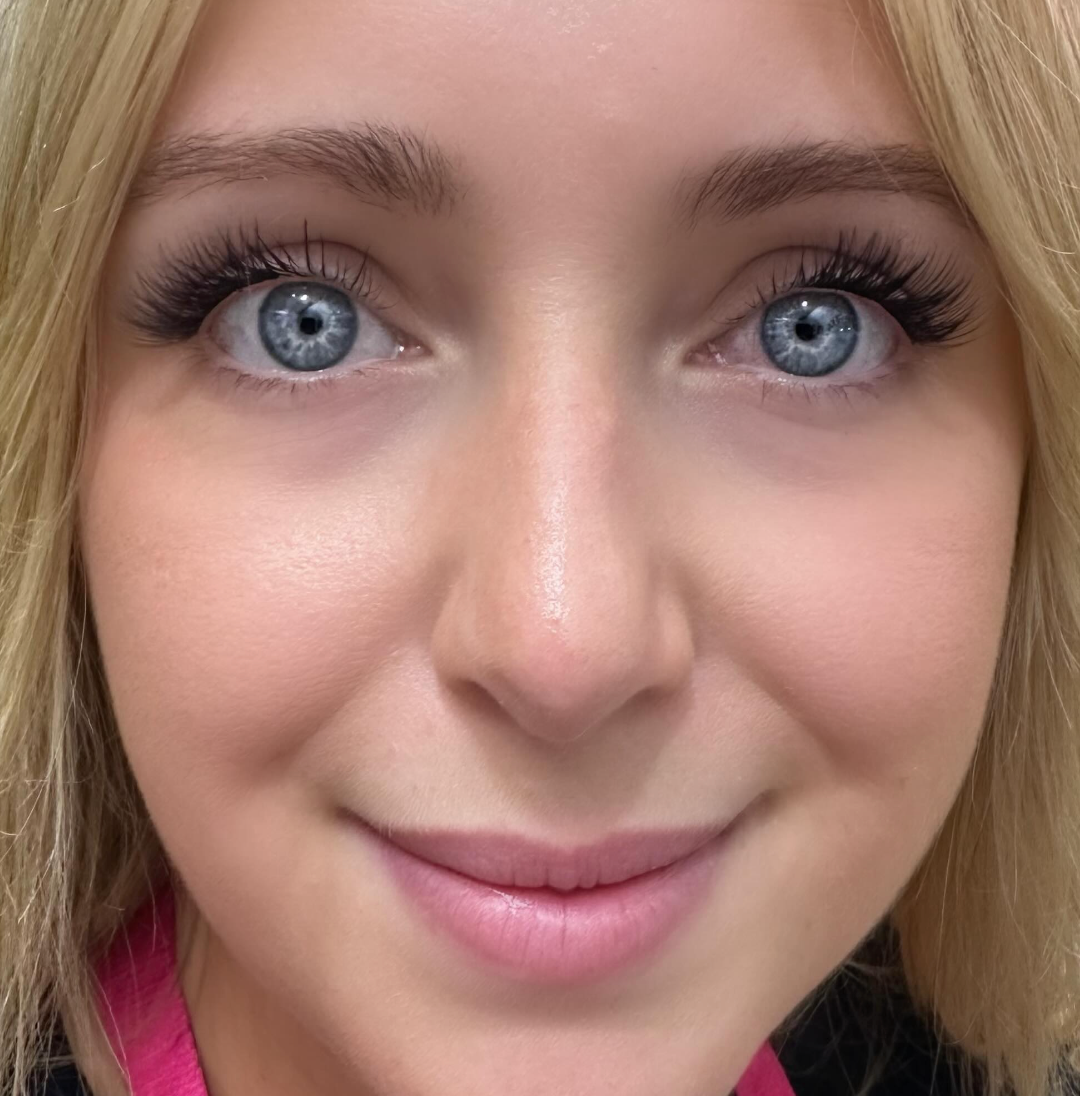 A person with blue eyes and light blonde hair smiles in a cozy home studio while showcasing their natural beauty without needing the Lash Link DIY Eyelash Extension Kit. They&
