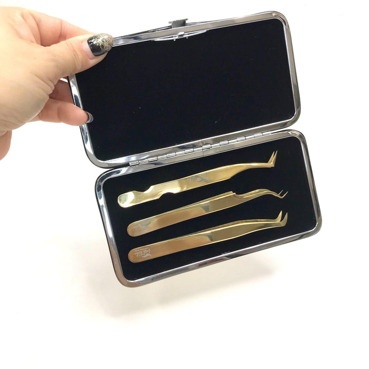 A person holds a Gold Magnetic Tweezer Case by Lash Tribe, featuring an open rectangular design in black or gold and containing three gold metallic tweezers with different tip designs.