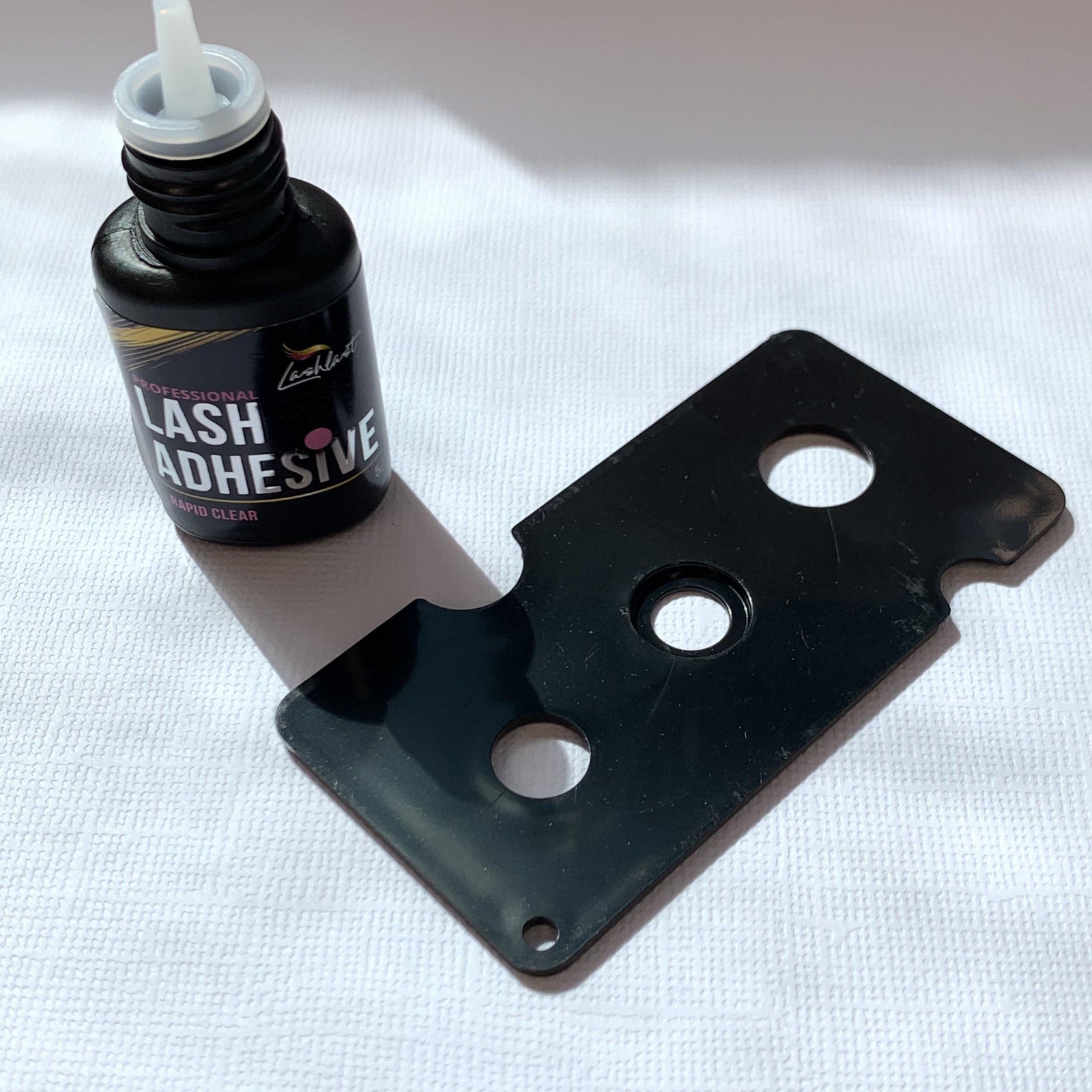 A bottle of Lash Tribe Lash Glue Nozzle Opener next to a black piece of plastic.