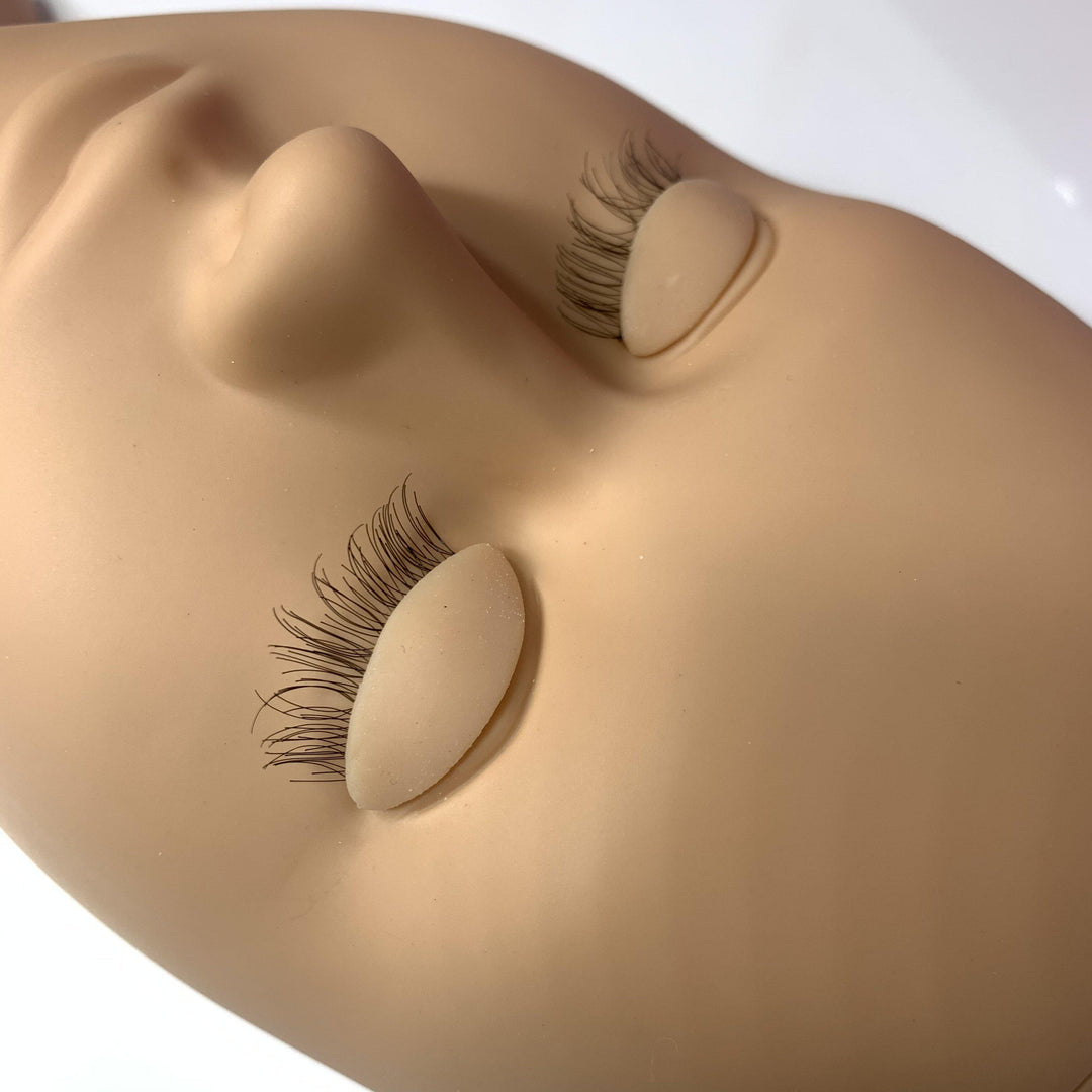A close-up of the Lash Tribe Mannequin Practice Head with closed eyes, featuring long, curly eyelashes applied to both interchangeable eyelids.